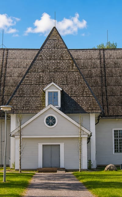 Elimaki's large white wooden church with a steep shingled roof and birch-lined path under a blue sky.