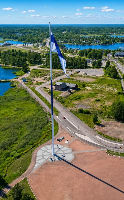 Aerial view of Hamina, Finland, with a giant Finnish flag over a circular waterfront plaza, marina and Gulf archipelago.