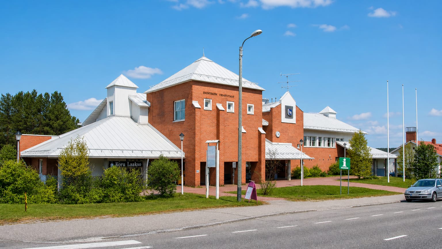 The red-brick Enontekioe municipal office building with white roofs beside a roadside on a clear summer day.
