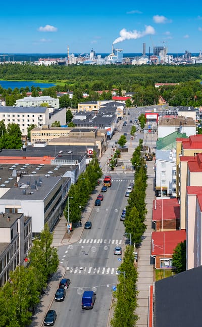 Aerial view of central Kemi, Finland: a wide tree-lined street through low-rise blocks, with the pulp mill on the bay horizon.