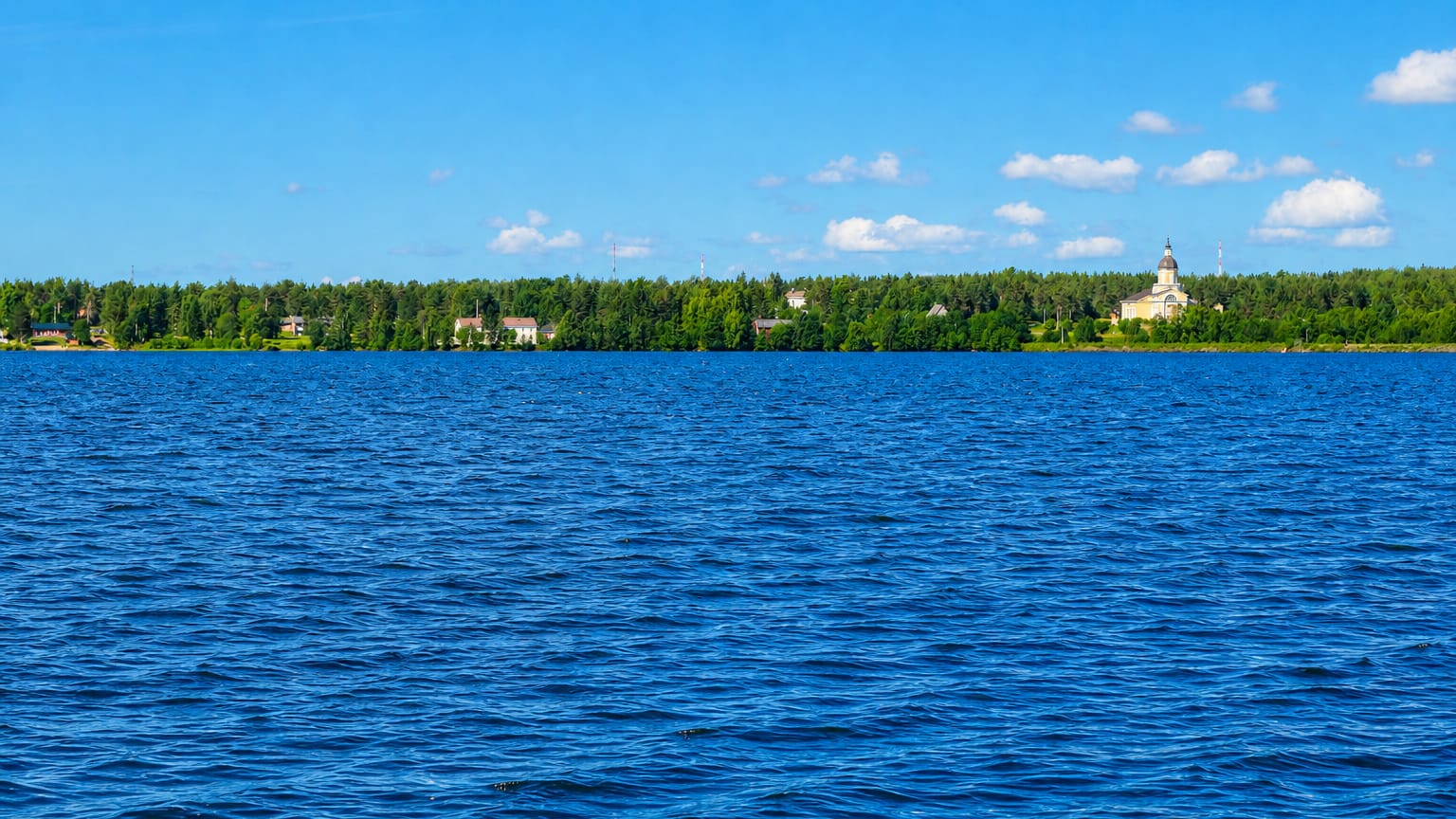Calm blue water leading to Keminmaa's forested shoreline and a distant white church tower under a bright summer sky.