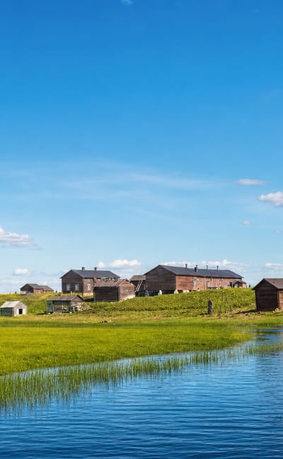 Traditional red wooden farm buildings on a green riverbank under a bright blue sky in Kittilä, Finnish Lapland.