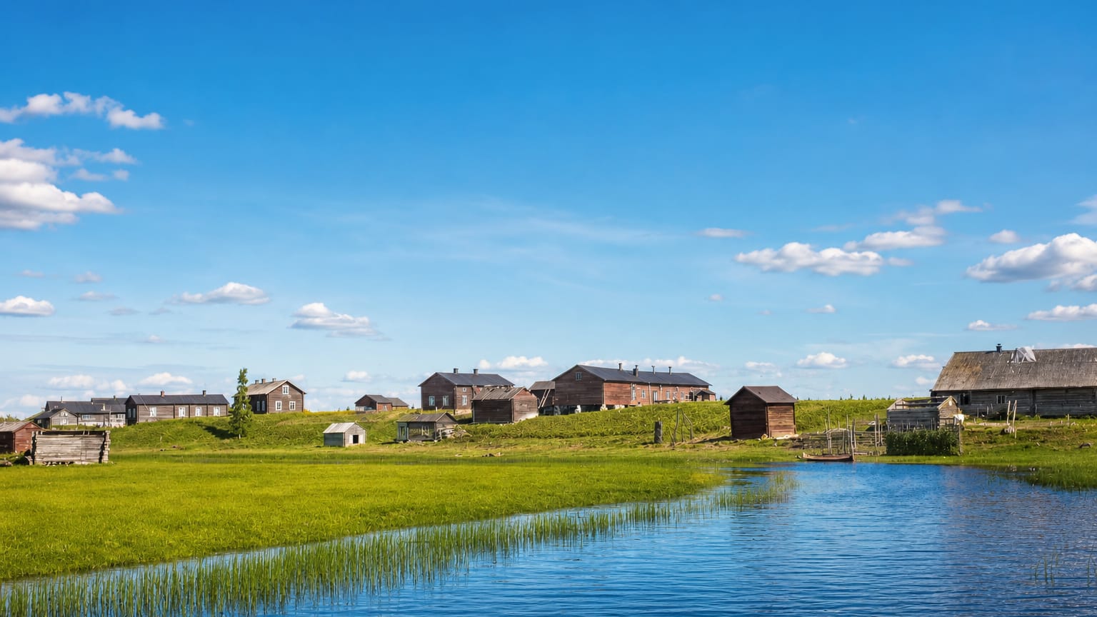Traditional red wooden farm buildings on a green riverbank under a bright blue sky in Kittilä, Finnish Lapland.