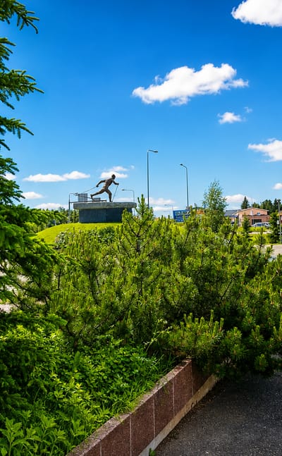 Sunny summer square in Pello, Finland, with a bronze runner statue, pine trees and a red building beside an open paved lot.