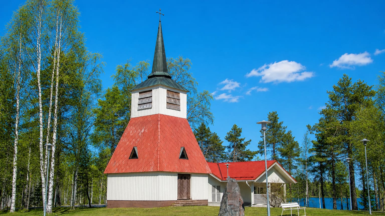 White and red wooden bell tower church beside a lake among birch trees in Posio, Finland.