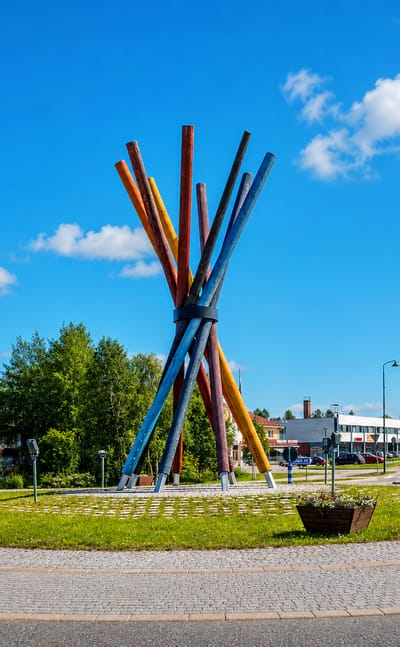 Colourful log-pile sculpture on a roundabout at the centre of Salla, Finland, with low commercial buildings behind.