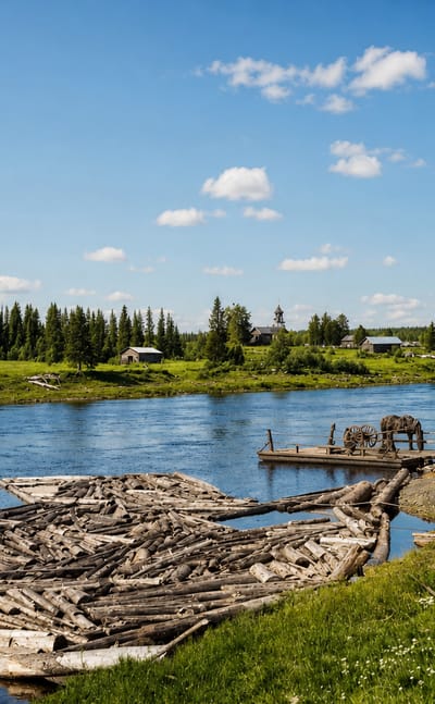 Log rafts float on a calm river beside grassy banks, spruce forest and scattered wooden cabins at Savukoski, Finland.