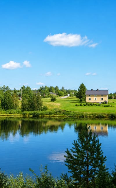 A calm river reflecting a yellow farmhouse and red barn among pine forest near Sodankylae in Finnish Lapland.