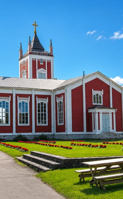 Tervola's red wooden church with white trim and a cross-topped tower above a flower-lined green churchyard.