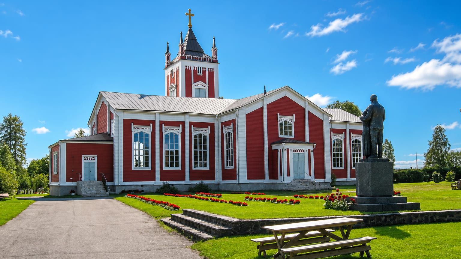 Tervola's red wooden church with white trim and a cross-topped tower above a flower-lined green churchyard.