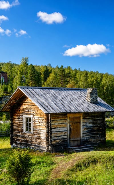 Wooden log church huts in a green meadow below Utsjoki's hilltop church, framed by Lapland forest under a blue sky.