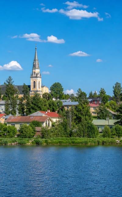 Lakeside view of Artjärvi, Finland, with a yellow church spire and a water tower rising above wooden houses and trees.