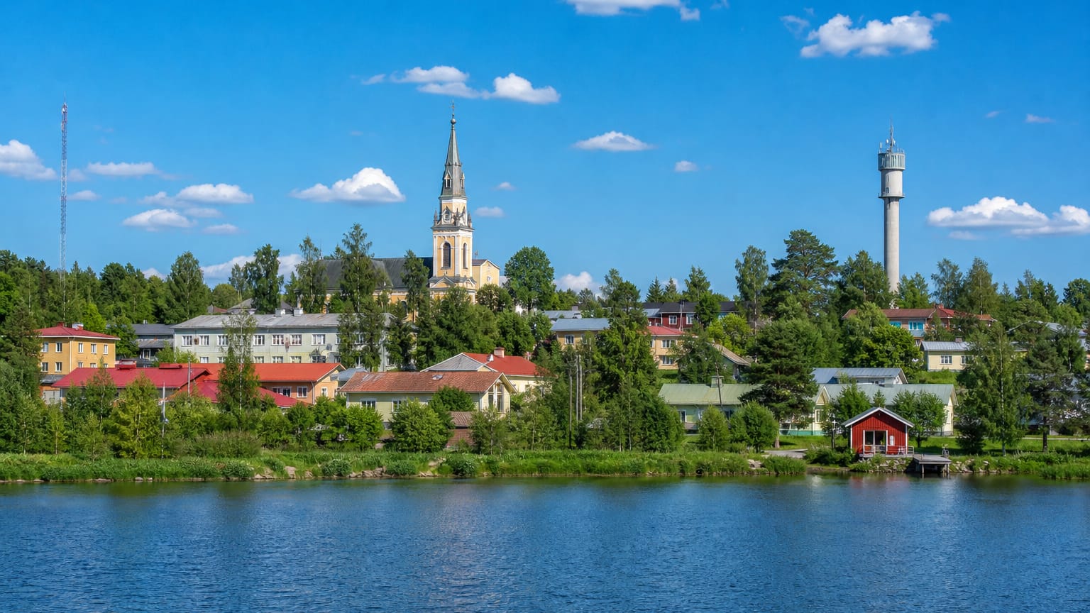 Lakeside view of Artjärvi, Finland, with a yellow church spire and a water tower rising above wooden houses and trees.
