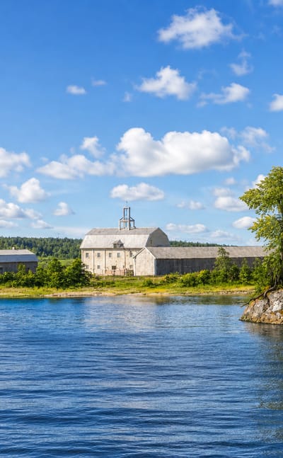 Heinola, Finland: calm river framed by rocky pine and birch shores with pale waterfront buildings under a blue summer sky