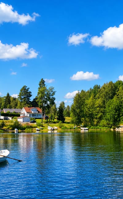 A person rows a small boat across a calm lake at Ikaalinen, Finland, past a forested shore with cottages and moored boats.