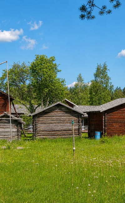 Traditional red-timber farm buildings and log storehouses in a green summer meadow in Juupajoki, Finland.