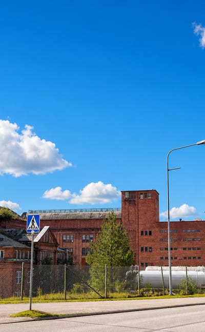 Mänttä's historic red-brick Serlachius pulp mill and tall smokestack beside a roadside under a clear blue summer sky.