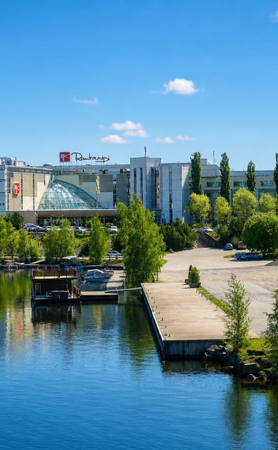 Sunny waterfront in Nokia, Finland: a lakeside spa hotel and marina with moored boats along a calm, forested shore.