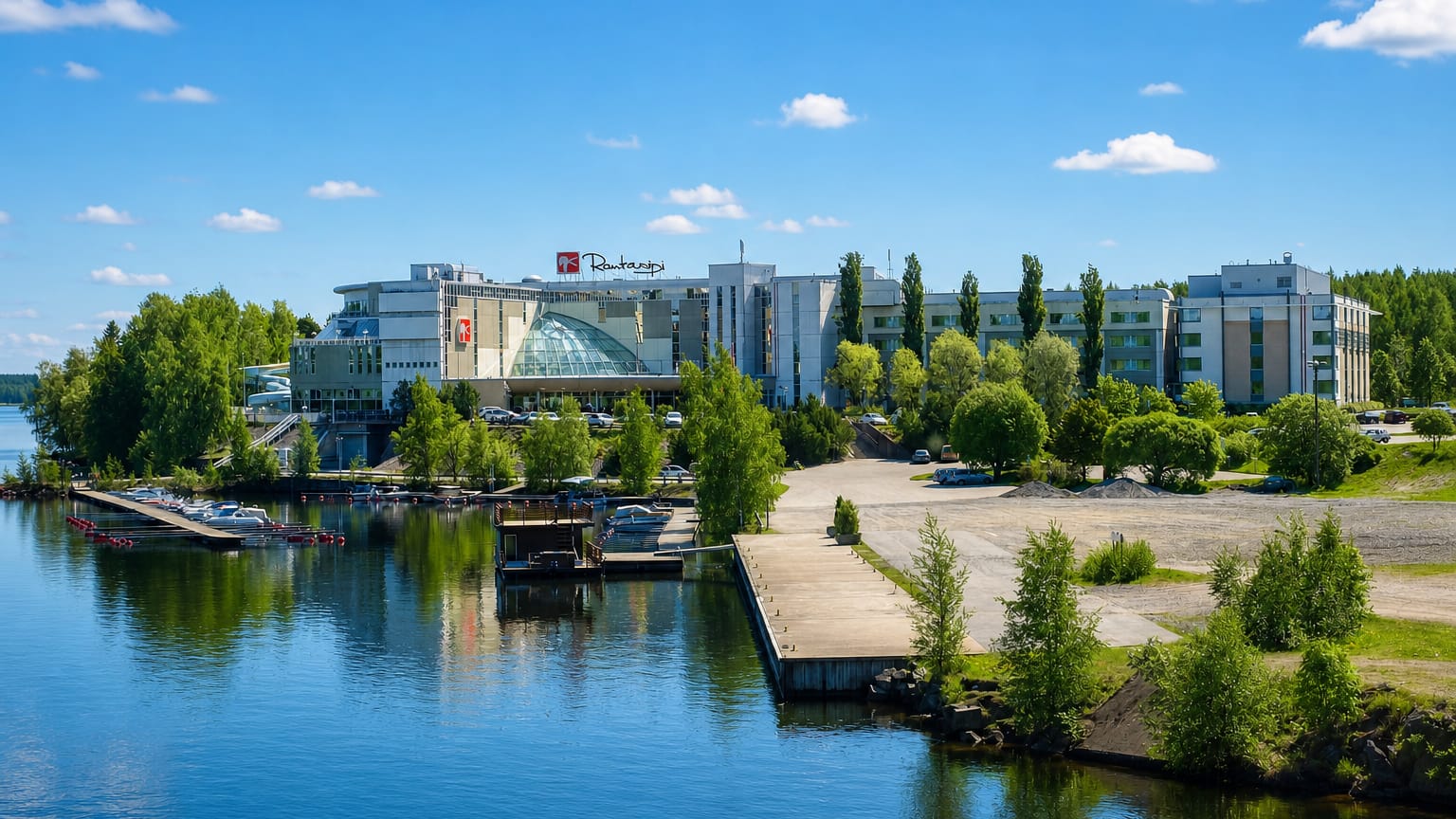 Sunny waterfront in Nokia, Finland: a lakeside spa hotel and marina with moored boats along a calm, forested shore.