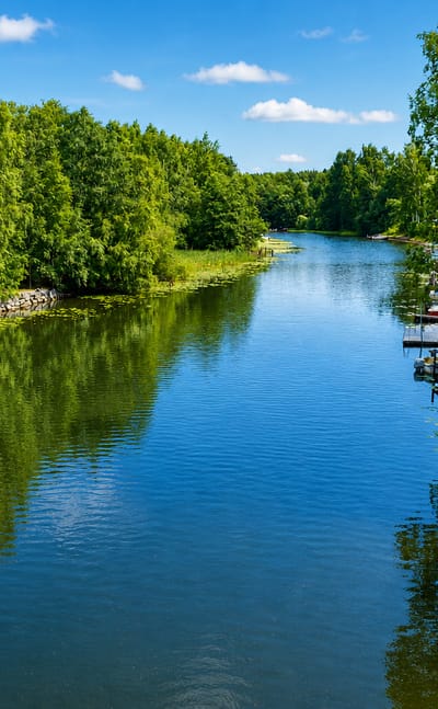 Calm tree-lined waterway in Pälkäne, Finland, with a lakeside boathouse and small boats moored at wooden docks under a blue sky.