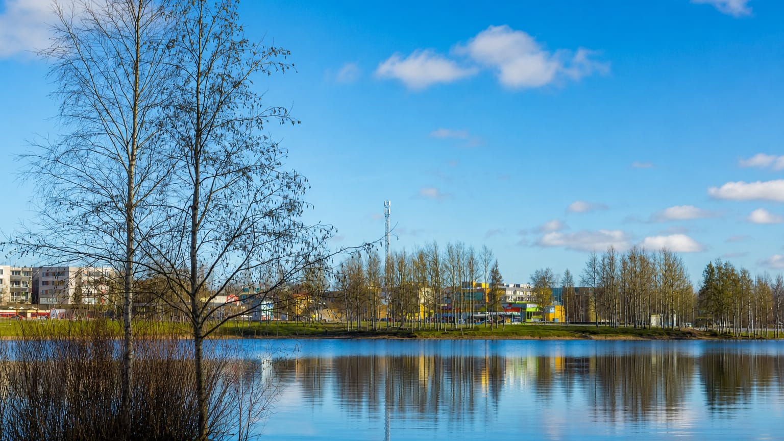 Calm lake reflecting Parkano's low-rise apartment and commercial buildings beneath a blue Finnish spring sky.