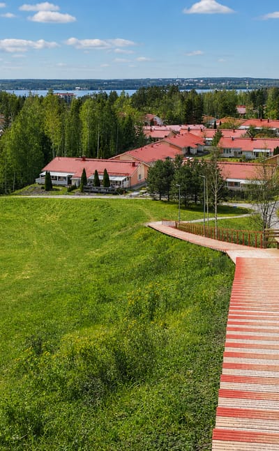 Red wooden staircase descending a green hillside toward red-roofed houses and a lake in Pirkkala, Finland on a sunny day.