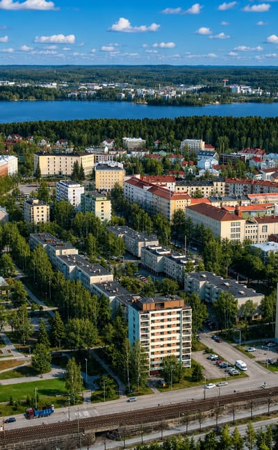 Aerial daytime view of Amuri, Tampere, apartment blocks and leafy streets beside a blue lake under a cloud-dotted sky.