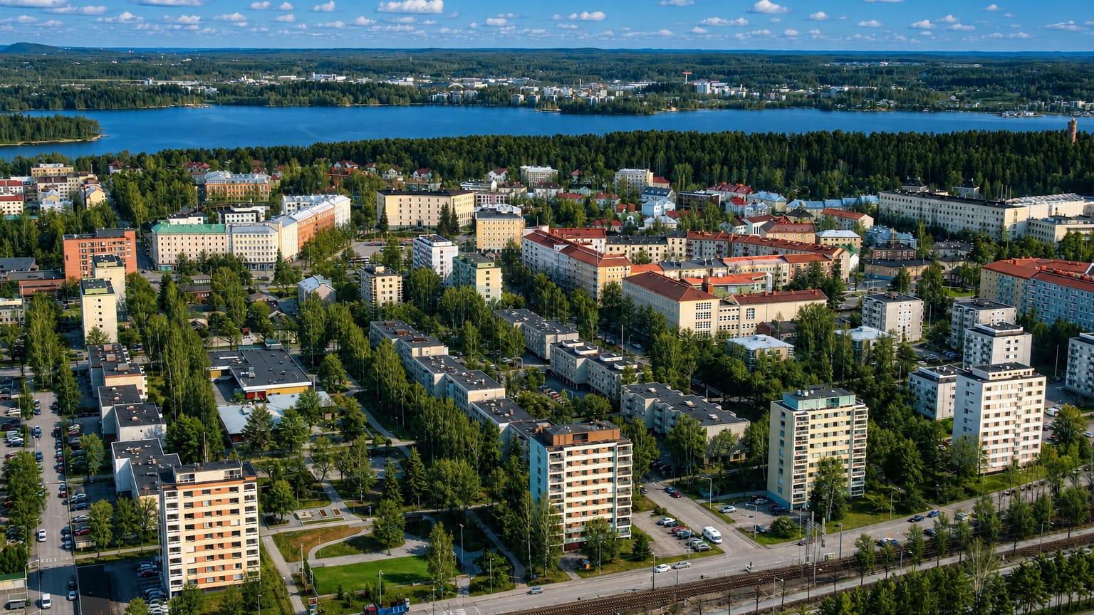Aerial daytime view of Amuri, Tampere, apartment blocks and leafy streets beside a blue lake under a cloud-dotted sky.