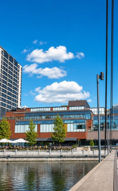 Cyclists cross a bridge over the Tammerkoski in Kyttälä, Tampere, past a riverside tower and glass-domed mall.