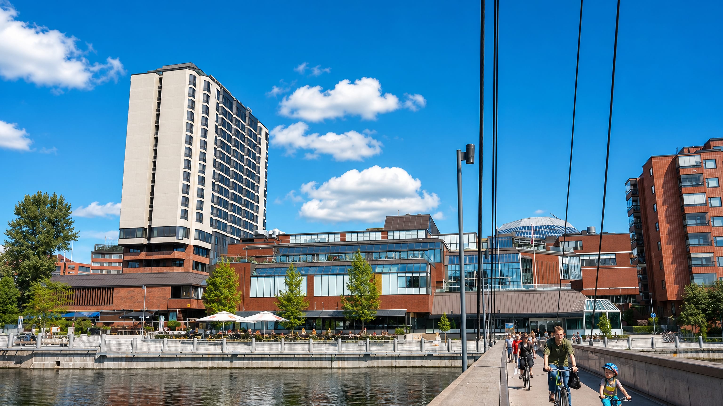 Cyclists cross a bridge over the Tammerkoski in Kyttälä, Tampere, past a riverside tower and glass-domed mall.