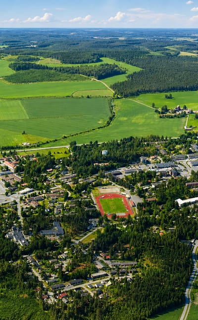 Aerial view of Urjala town centre in Pirkanmaa, Finland, with its sports field, low buildings, farmland, forest and a small lake under a summer sky.