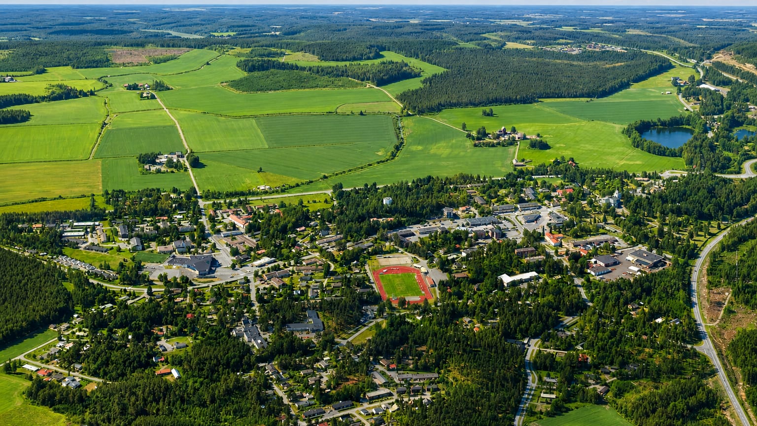 Aerial view of Urjala town centre in Pirkanmaa, Finland, with its sports field, low buildings, farmland, forest and a small lake under a summer sky.