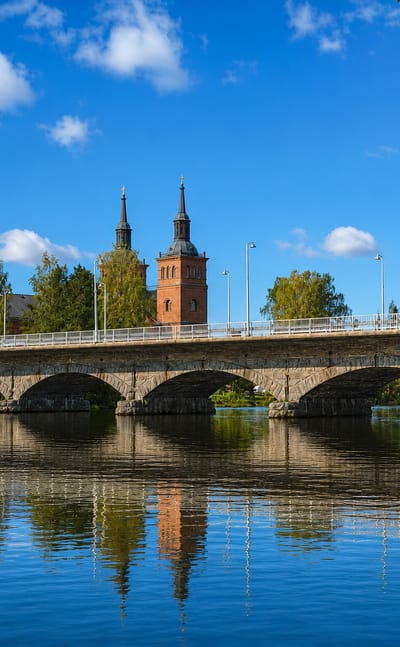 Stone arch bridge over calm water in Vammala, Finland, with the red-brick twin-towered town church beyond autumn trees