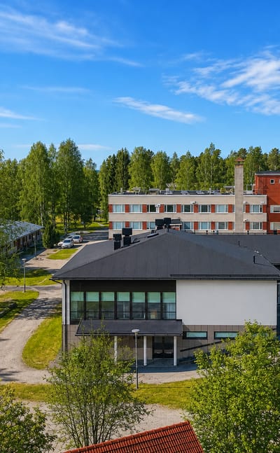 Aerial view of Virrat, Finland, showing a multi-storey building among birch trees with a forested horizon under clear blue sky.