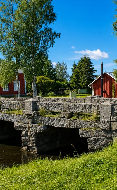 Old stone-arch bridge over a stream beside red wooden houses and birches in Korsnäs, Finland.