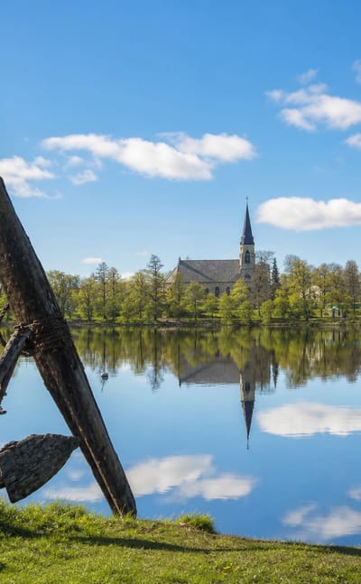 Kristinestad waterfront with the town church and spire reflected in calm water and an old wooden anchor on the grassy shore