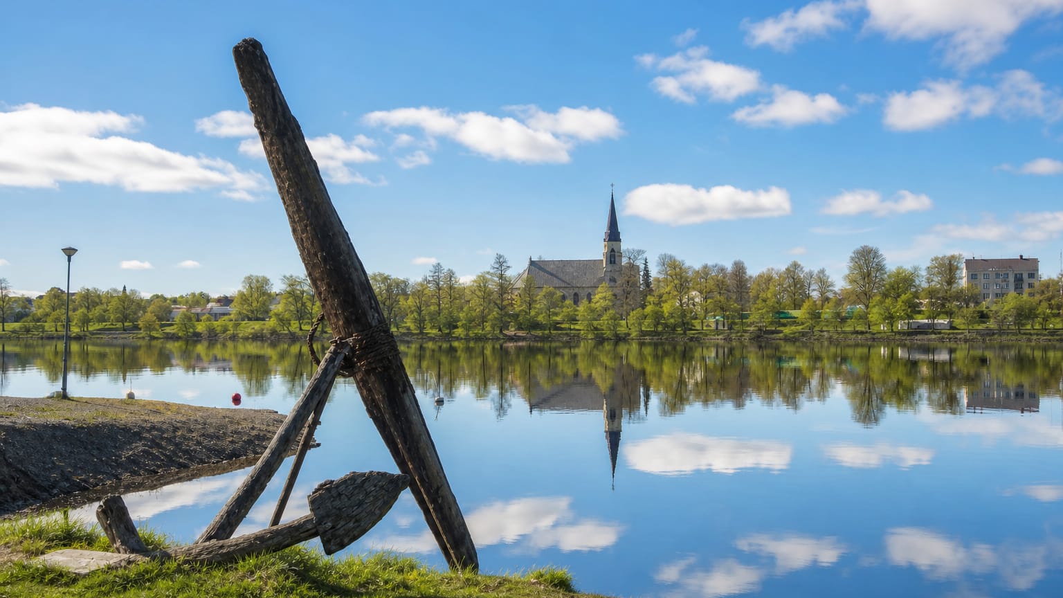 Kristinestad waterfront with the town church and spire reflected in calm water and an old wooden anchor on the grassy shore
