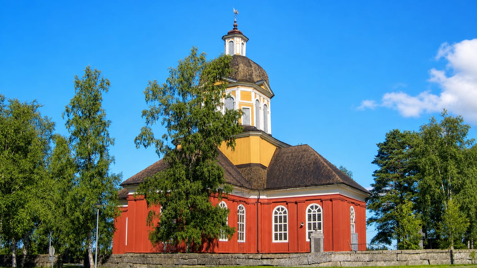 Red wooden church with an octagonal domed roof and yellow lantern tower behind a stone wall in Larsmo, Finland.