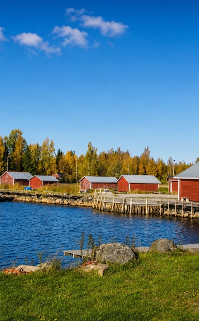 Red wooden boathouses and small boats lining a calm inlet at Malax, Finland, amid autumn trees.