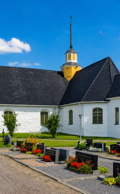 White stone church with two yellow-domed towers and a graveyard in Narpes, Finland, under a blue sky.