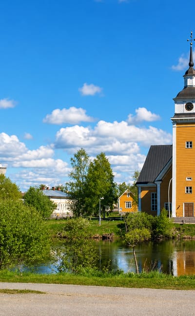 Nykarleby's yellow wooden church and freestanding bell tower reflected in the riverside under a blue summer sky.