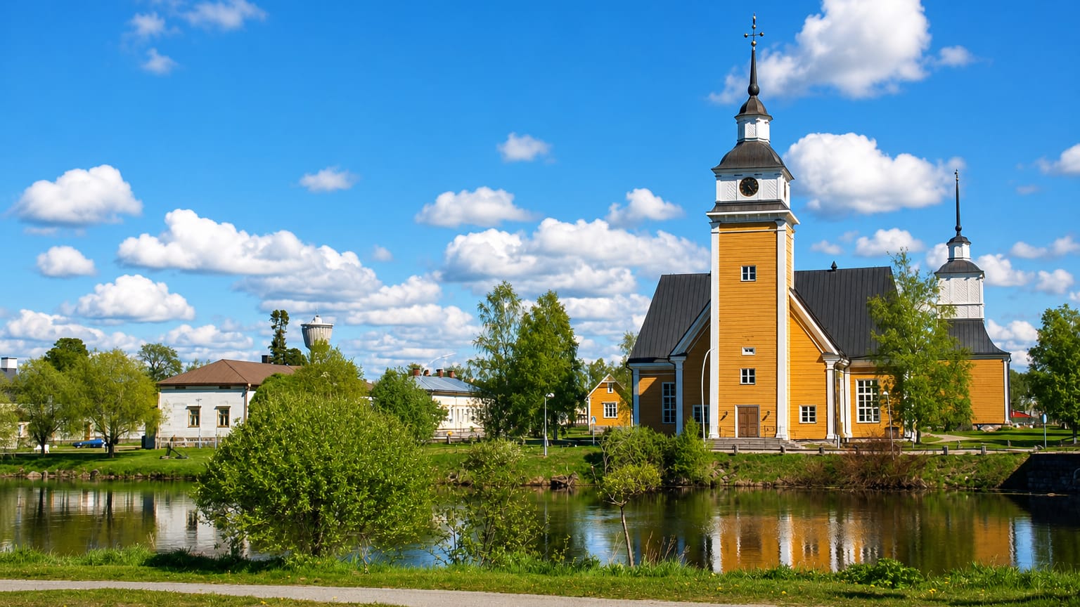 Nykarleby's yellow wooden church and freestanding bell tower reflected in the riverside under a blue summer sky.