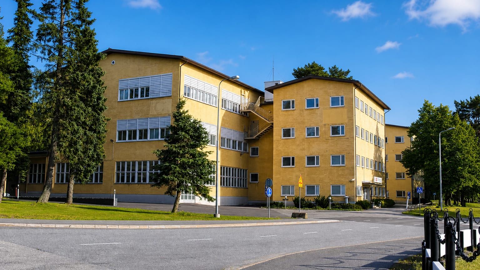 Yellow functionalist institutional building among pines on a quiet sunlit street in Huutoniemi, Vaasa.