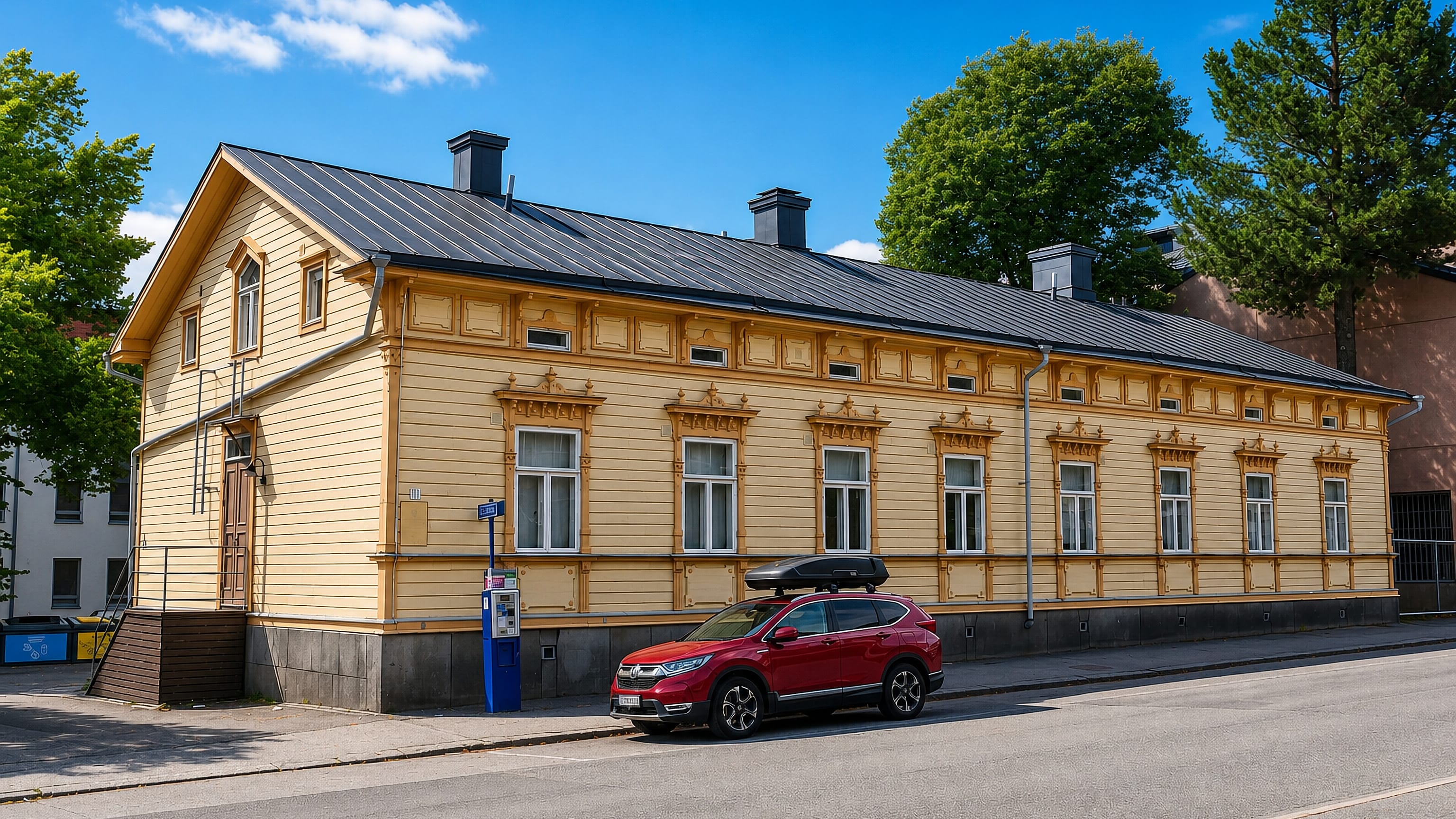 Ornate yellow wooden house with a seam-metal roof along a sunny street in III kaupunginosa, Vaasa.
