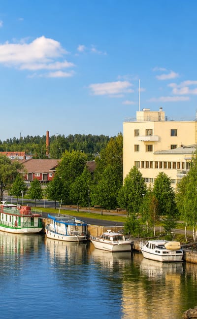 Small boats moored along a calm river quay in Joensuu, Finland, below a pale building and forested green banks