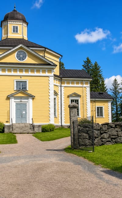 Juuka's yellow neoclassical wooden church with a clock and domed cupola behind a stone wall and gravel path.