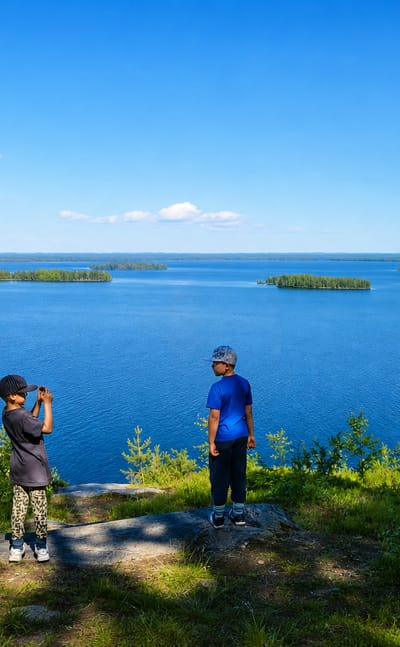 Two children on a rocky viewpoint overlooking a forested lake with islands near Polvijärvi in summer.