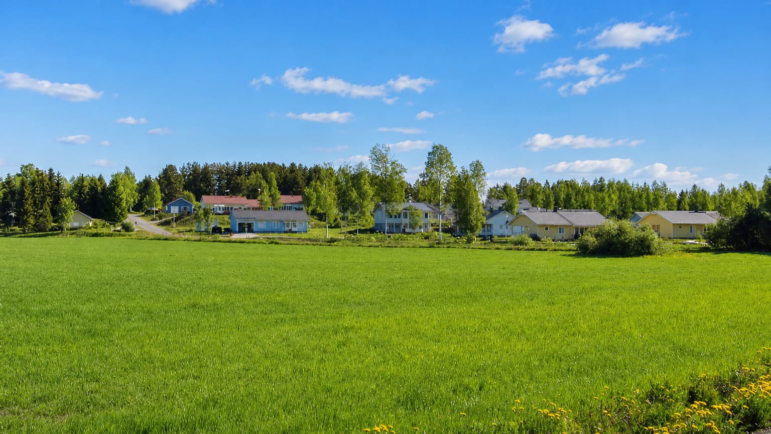 Wooden houses of Pyhäselkä village behind a bright green summer meadow with forest and blue sky, North Karelia, Finland