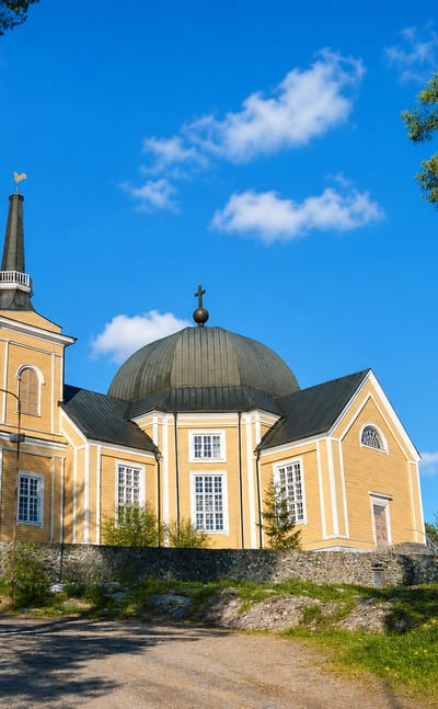 Yellow wooden church with a domed roof and bell tower on a hilltop in Rääkkylä, Finland, under a blue sky.