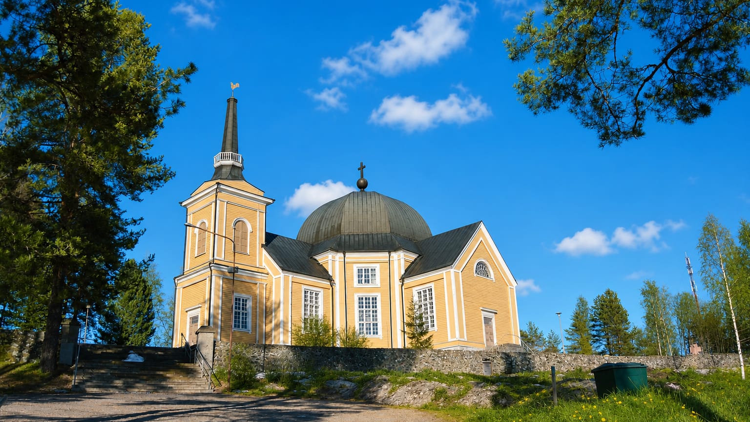 Yellow wooden church with a domed roof and bell tower on a hilltop in Rääkkylä, Finland, under a blue sky.